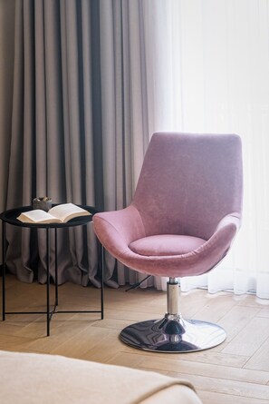 Ein modernes Zimmer mit einem rosafarbenen Sessel, einem kleinen schwarzen Beistelltisch mit einem offenen Buch und einem großen Fenster mit Vorhängen.