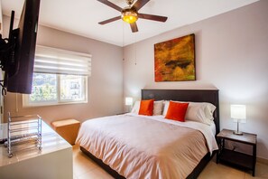 A hotel room with a bed, a ceiling fan, a window with blinds, a bedside table with a lamp, and a TV mounted on the wall.
