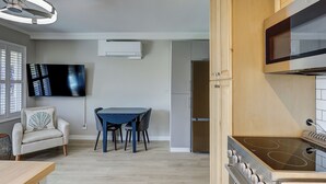 Een moderne keuken met een inbouwoven, een eettafel met stoelen, een aan de muur bevestigde televisie en een raam met jaloezieën.