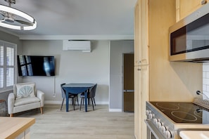 A modern kitchen with a built-in oven, a dining table with chairs, a TV mounted on the wall, and a window with blinds.