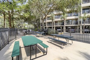 Un patio al aire libre con mesas de picnic y bancos, rodeado por una valla metálica, con un edificio de varios pisos al fondo.