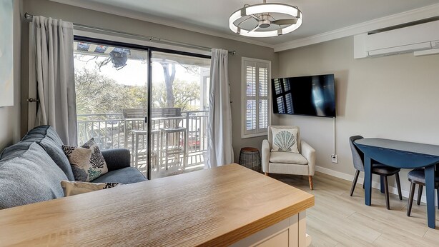 Een woonkamer met een bank, een houten tafel, een aan de muur bevestigde televisie en een balkon met uitzicht op bomen.