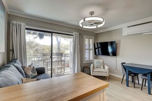 A living room with a sofa, a wooden table, a TV mounted on the wall, and a balcony with a view of trees.