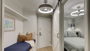 Een smalle gang die leidt naar een slaapkamer met een bed, een nachtkastje en een lamp.