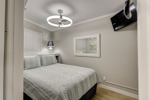 A bedroom with a bed, a ceiling fan, a TV mounted on the wall, a window with blinds, and a bedside table with a lamp.