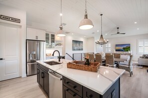 A modern kitchen with a large island, stainless steel appliances, and a dining area with wicker furniture and a TV. - Private Pool! Ping-Pong, Pool Table, Putt-Putt! (Holmes Beach)