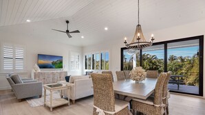 A spacious living room with a dining area, large windows with shutters, a ceiling fan, and a chandelier.