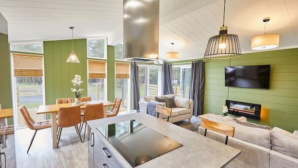 A modern kitchen with a central island, dining area with chairs, a sofa, and a TV mounted on the wall. - Host & Stay | Eller Beck - The Cabin in the Woods (Whitby)