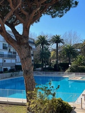 Une piscine entourée d’une clôture, avec un arbre au premier plan et un bâtiment en arrière-plan.