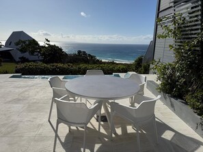An outdoor patio with a round table and white chairs, overlooking a beach and ocean. - Luxury, magnificent views, space and swimming pool (Point Lookout)