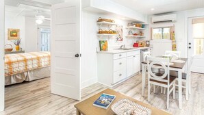 Un interior luminoso y blanco con una zona de cocina que incluye armarios blancos, una mesa de comedor con sillas y una puerta visible que da a otra habitación.