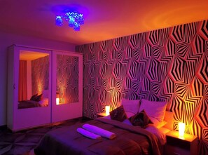 Une chambre avec un papier peint à motifs, un lit avec des oreillers, une table de chevet avec une lampe et une porte coulissante dotée d’un miroir.