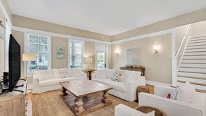 A spacious living room with a large white sofa, a wooden coffee table, and a television on a wooden stand. There are two windows with blinds, a lamp on a side table, and a staircase to the right. - Gorgeous Watercolor Home Close to  Camp with NEW 6  "LSV" Low Speed Vehicle. (SANTA ROSA BEACH)