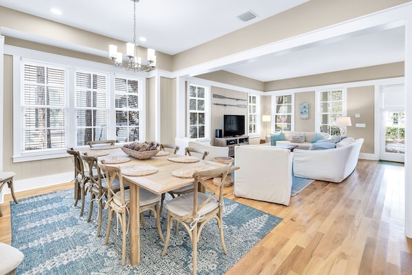 A spacious living room with a large TV, white sofas, and a dining area with a wooden table and chairs.