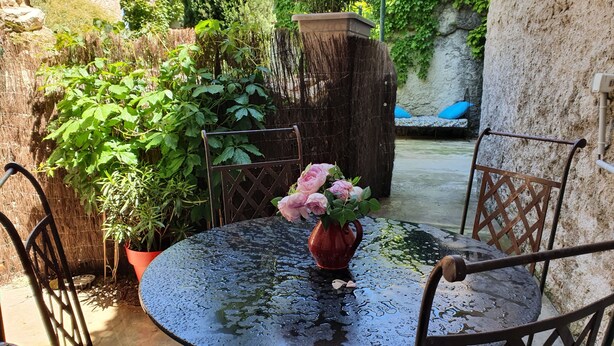 A round table with a vase of pink flowers, surrounded by metal chairs, in a garden with various plants and a stone wall. - House (Rochefort-du-Gard)