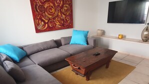 Een woonkamer met een grijze bank, blauwe kussens, een houten salontafel, een abstract schilderij in rood, een televisie en een klein rekje met decoratieve objecten.