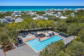 A resort with a large swimming pool surrounded by lounge chairs and umbrellas, set against a backdrop of ocean and beach houses.