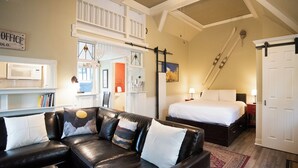 Ein Hotelzimmer mit schwarzem Ledersofa, einem Bett mit weißer Bettwäsche, Holzboden und einer weißen Tür.