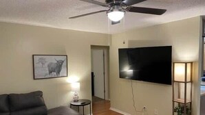 Een woonkamer met een bank, een plafondventilator, een flatscreen-tv, een lamp en een ingelijst schilderij aan de muur.