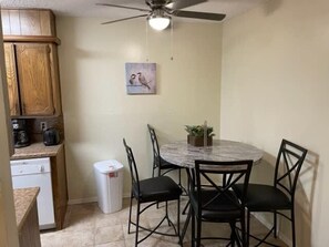Une cuisine compacte avec des meubles en bois, un lave-vaisselle blanc, une table à manger en marbre avec quatre chaises, un ventilateur de plafond et une petite plante en pot.