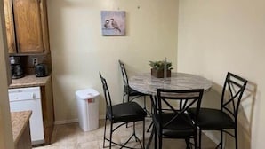 Een compacte keuken met houten kastjes, een witte vaatwasser, een marmeren eettafel met vier stoelen, een plafondventilator en een kleine potplant.