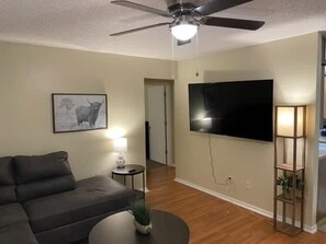 Un salon comprenant un canapé, un ventilateur de plafond, un téléviseur à écran plat, une lampe et un tableau encadré accroché au mur.