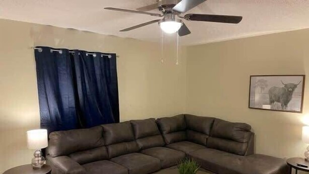Een woonkamer met een hoekbank, een plafondventilator, een kleine ronde tafel met een plant, en een lijst met een foto aan de muur.