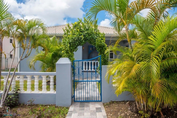 A gated entrance with a blue gate, a white balustrade, and lush greenery including palm trees.