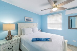 A bedroom with a bed, a ceiling fan, a nightstand with a lamp, a window with blinds, and a wall-mounted mirror.