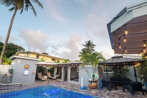 A pool area with a covered seating area, lounge chairs, and a pergola with string lights.