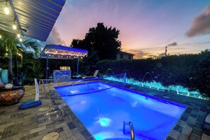 Una piscina con un borde iluminado en azul, una zona de estar cubierta y vistas al atardecer.