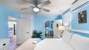 A bedroom with a bed, a ceiling fan, an air conditioning unit, a nightstand, a lamp, and a view of the bathroom.