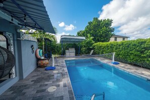 A swimming pool with a netted area, surrounded by a patio with stone tiles, a covered seating area, and lush greenery. - Private Pool | 3 Mins to Welches Beach | BBQ (Oistins)