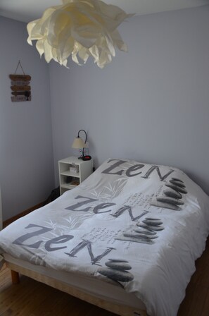 A bedroom with a bed covered in a white blanket with 'Zen' printed on it, a bedside table with a lamp, and a wall decoration.