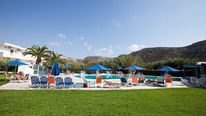 Zona de piscina con sillas de descanso y sombrillas, piscina y montañas al fondo.