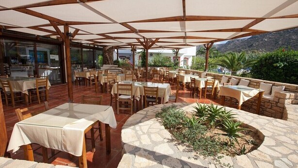 Un área de comedor al aire libre con mesas y sillas, una jardinera de piedra con plantas, y una vista de montañas al fondo.