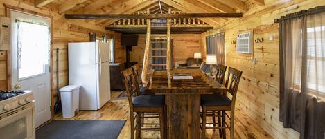 A wooden cabin interior with a dining table, chairs, a refrigerator, a stove, and a ladder leading to a loft.