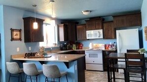 Una cocina con armarios de madera oscura, un refrigerador blanco, un microondas y una estufa. Hay dos taburetes de bar y una mesa de comedor con sillas.