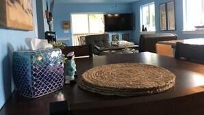 Una sala de estar con pared azul, ventilador de techo, televisor de pantalla plana, una mesa de centro y una mesa de comedor.