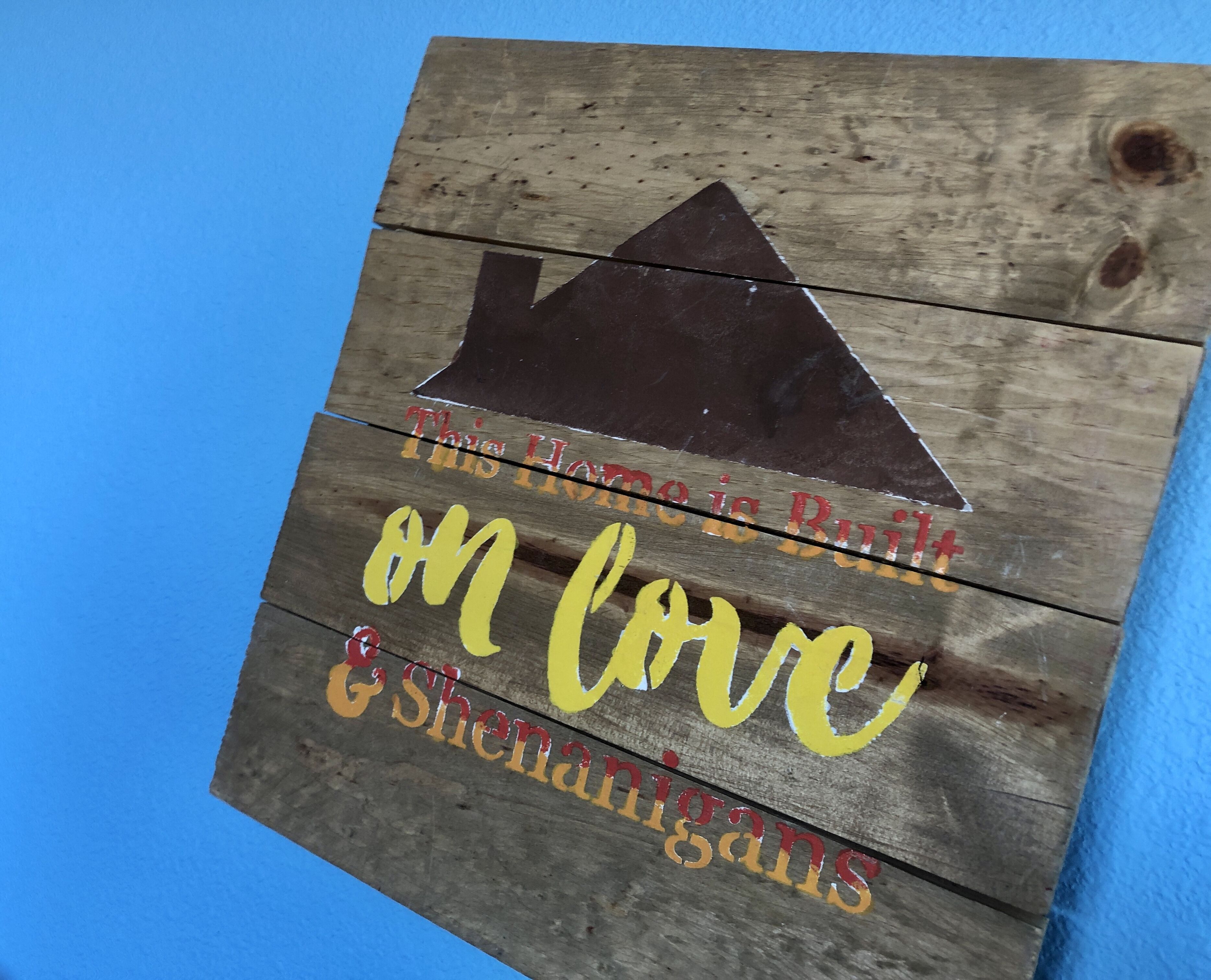 A wooden sign with the text "This Home is Built on Love & Shenanigans" and a triangle symbol.