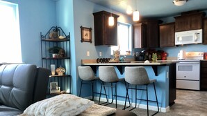 Una sala de estar con un sofá de cuero negro, un plaid blanco y un otoman gris. Hay tres taburetes grises en una isla de cocina con un microondas y una estufa. La cocina tiene armarios de madera oscura y luces colgantes.