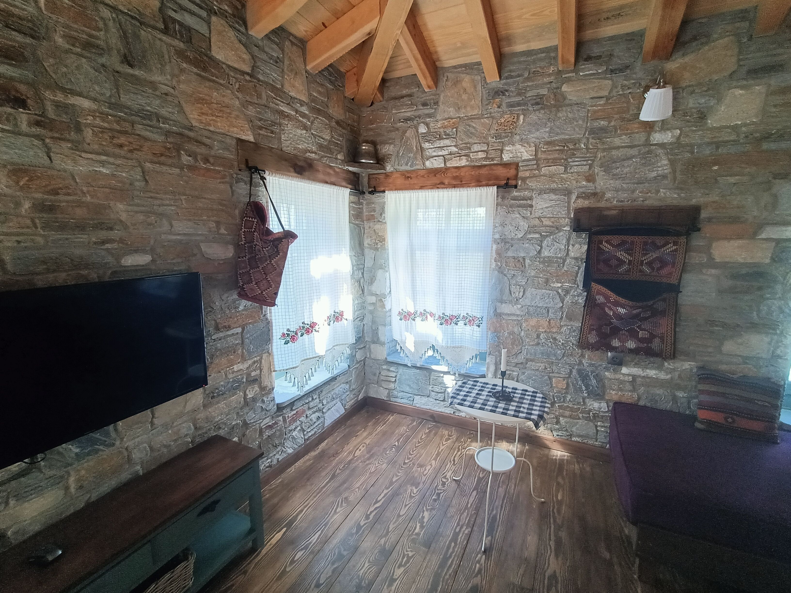 A stone-walled room with a wooden floor, a bench, a table with a checkered tablecloth, and a hanging bag.