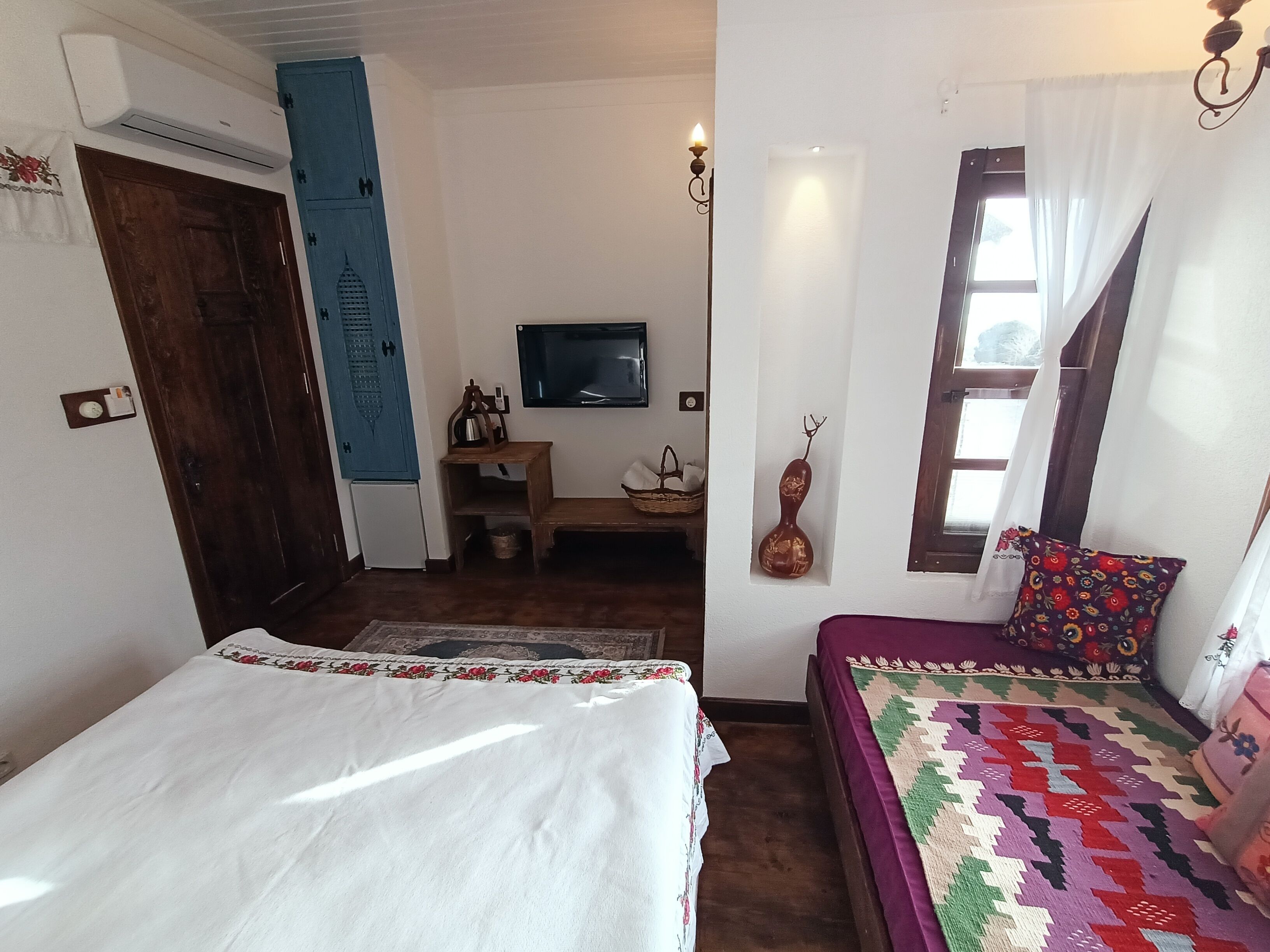 A bedroom with a bed, a TV, a small table, a wall-mounted shelf, and a window with curtains.