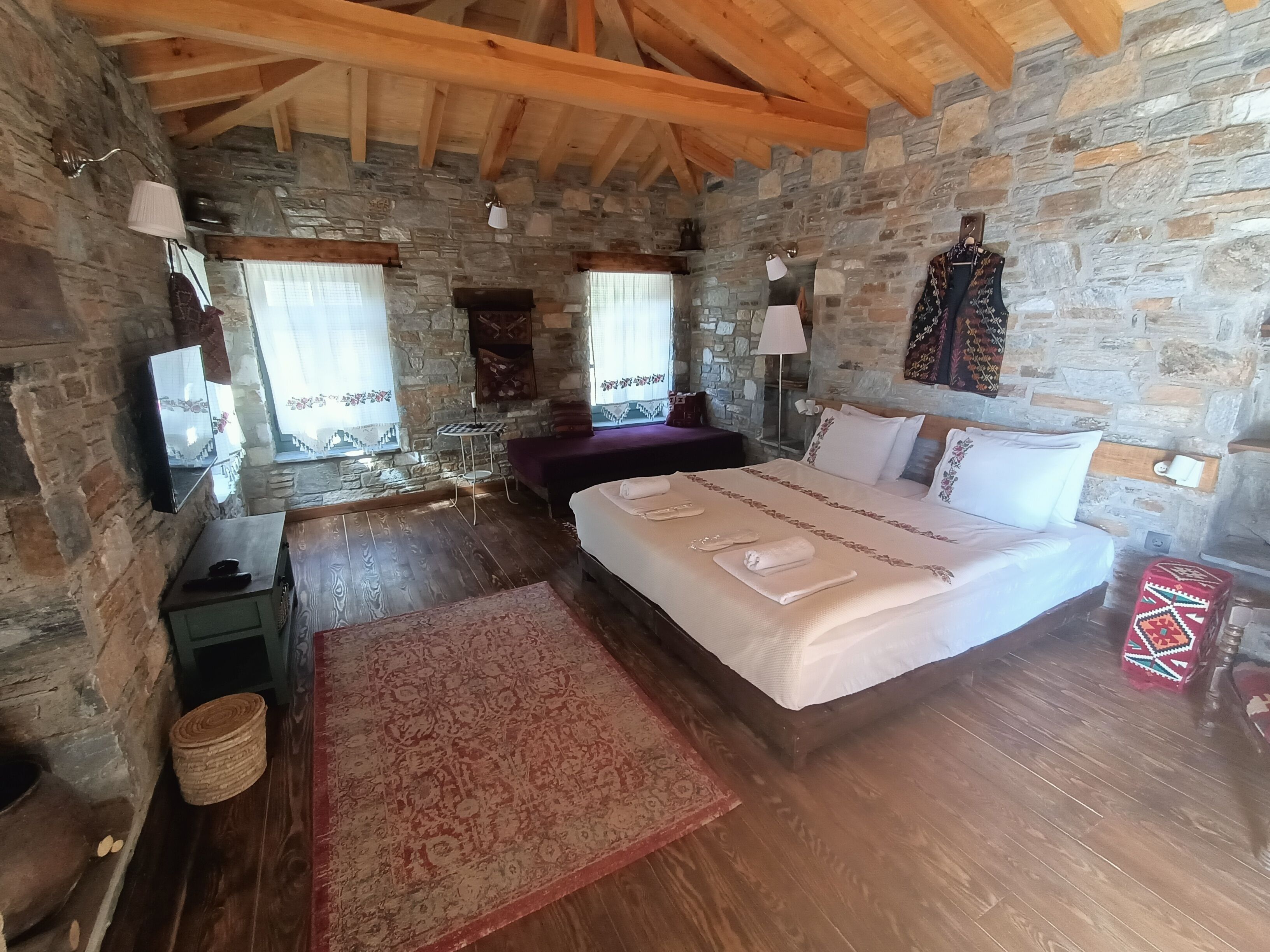 A rustic bedroom with stone walls, a wooden ceiling, a large bed, a nightstand, a bedside table, a lamp, a mirror, and a window with curtains.