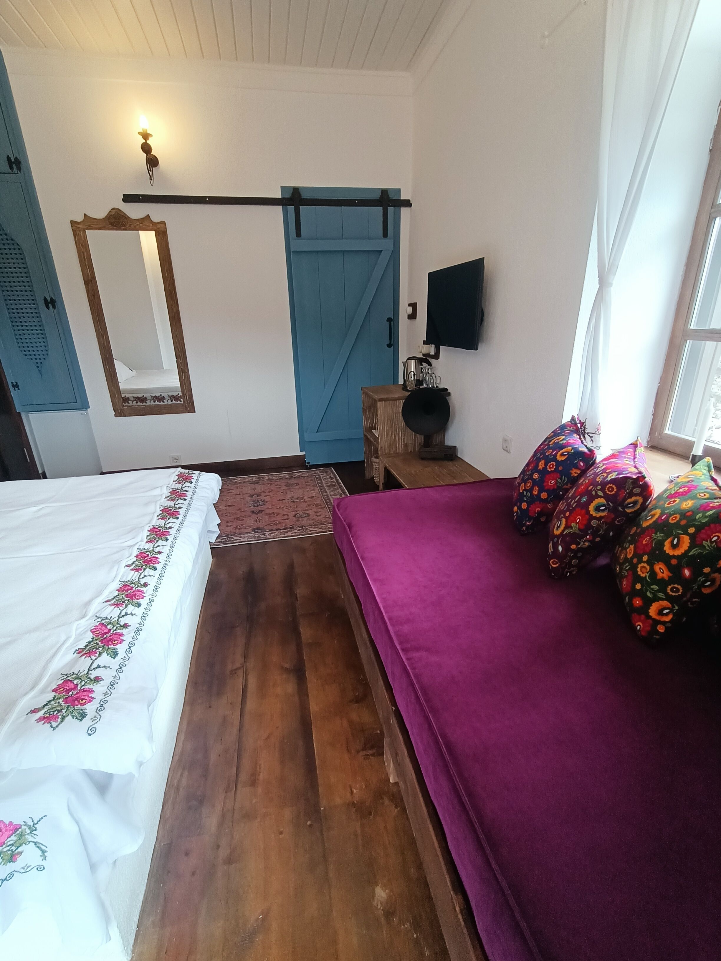 A bedroom with a wooden floor, a bed with a white bedspread, a purple sofa with colorful cushions, a blue door, a wall-mounted TV, and a mirror.