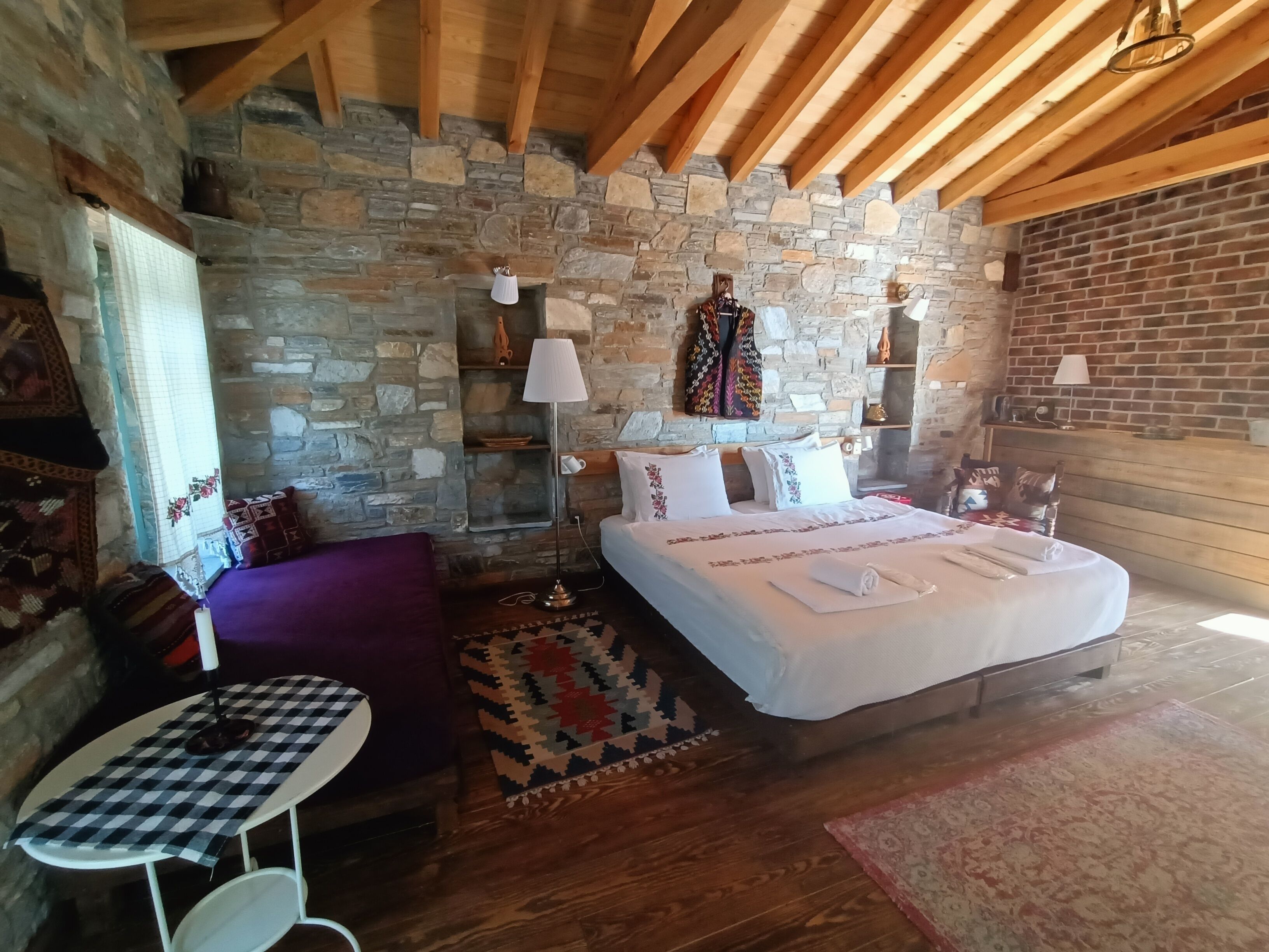 A cozy bedroom with a stone wall, wooden beams, a bed with white linens, a small table with a checkered tablecloth, and a purple sofa.