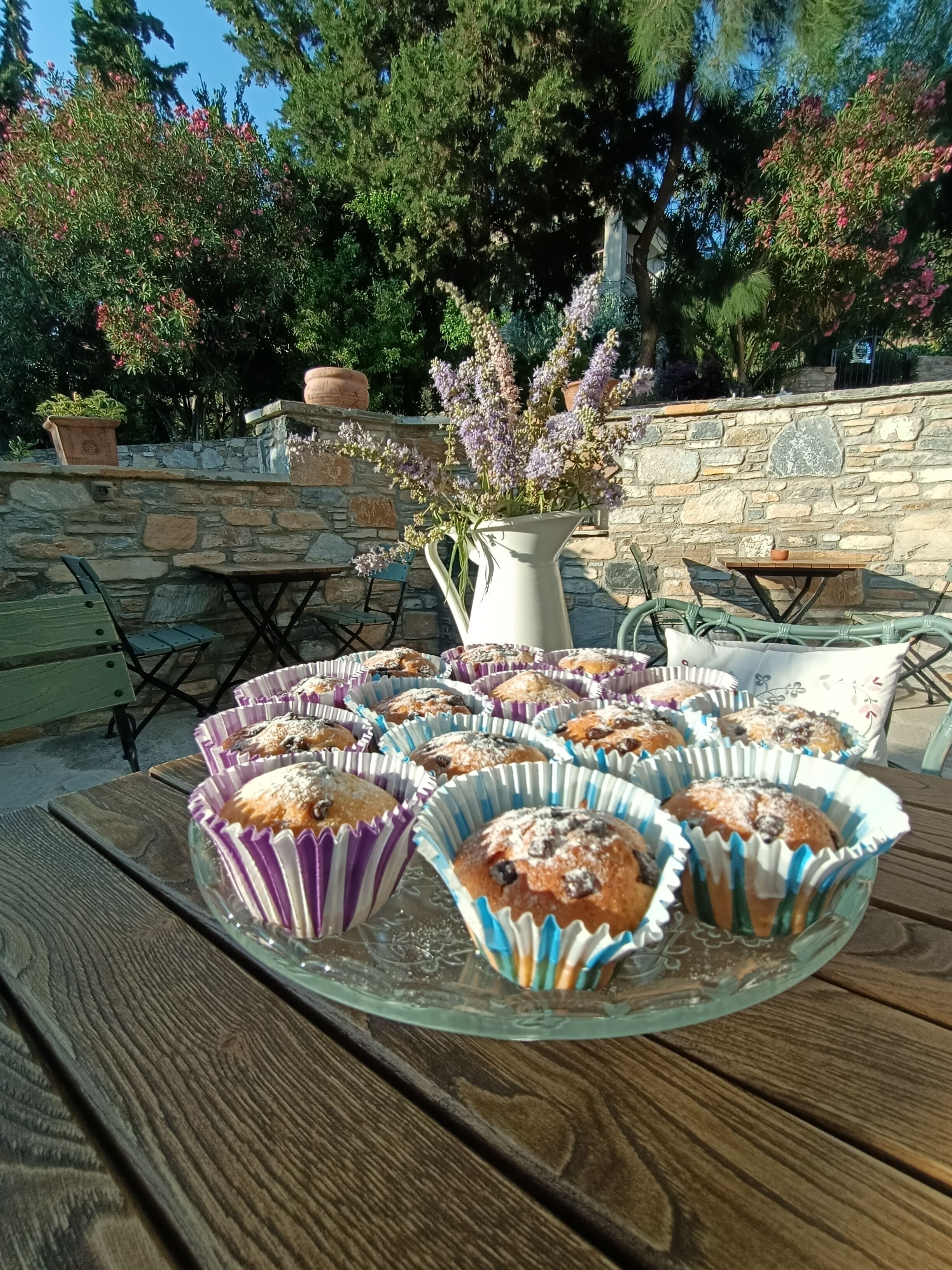 A table with a glass serving tray holding muffins, a white pitcher with flowers, and a stone wall in the background.