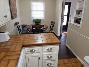 Uma cozinha com bancada em azulejos, uma torradeira, uma mesa de jantar com cadeiras, uma janela com persianas e uma porta que dá para outro cômodo.