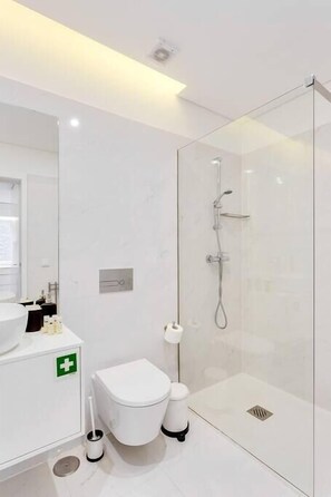 A modern bathroom with a glass shower enclosure, a white toilet, and a sink with a green emergency sign. - Almada Downtown Apartment by DA'Home (Porto)