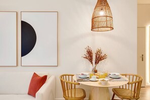 A dining area with a round table, two wooden chairs, a hanging light fixture, and wall art.